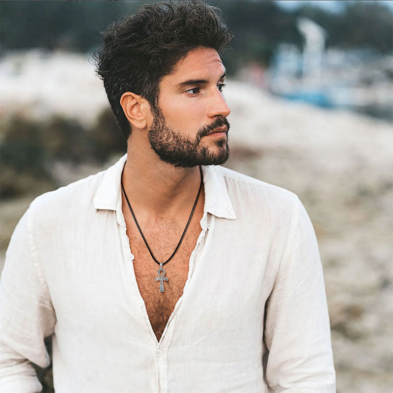 Homem de barba e cabelos ondulados usando o Colar Prata Masculino Ankh com cordão preto, camisa branca aberta e fundo praiano desfocado.