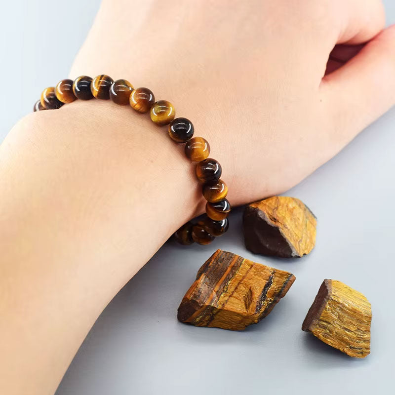 Pulseira Pedra Olho Tigre no pulso de uma pessoa, destacando suas contas arredondadas e brilho natural, com pedras brutas ao redor.